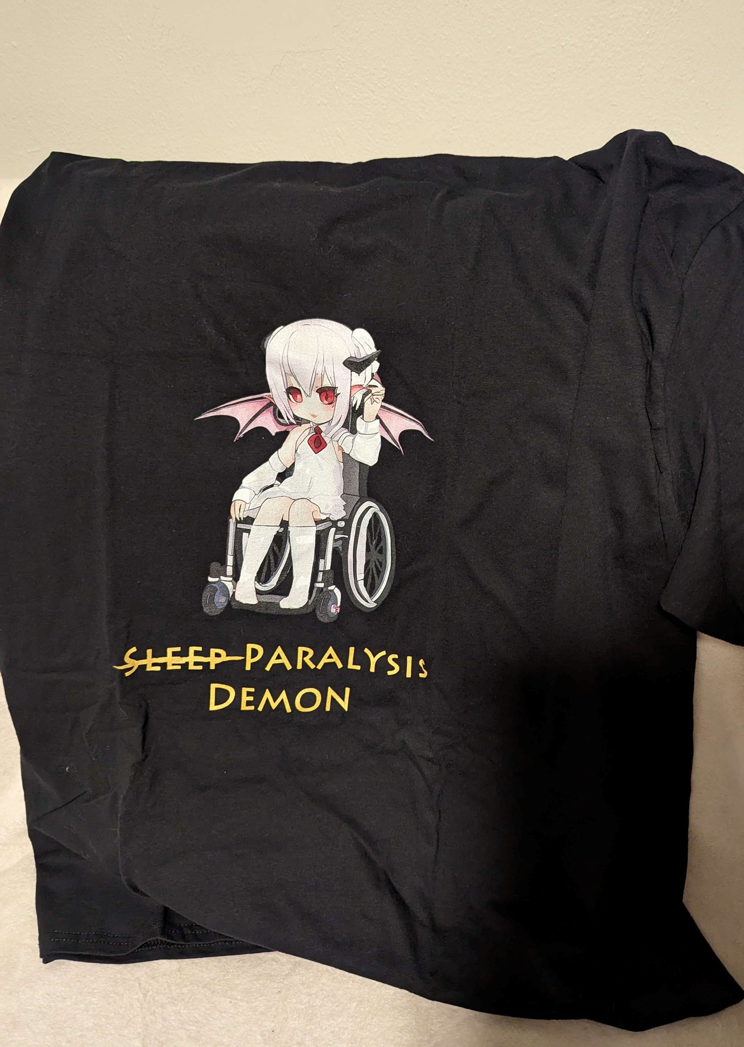 A black shirt featuring a graphic of a demon in a white shirt and skirt with a red tie sitting in a wheelchair. Below the graphic, there is gold text reading "Sleep paralysis demon" with sleep crossed out.