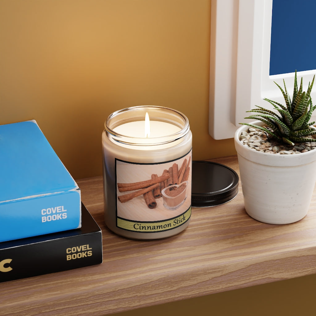 A lit cinnamon candle sitting on a shelf with books and a plant next to it.