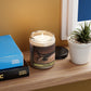 A lit vanilla candle sitting inbetween several books and a plant on a shelf.