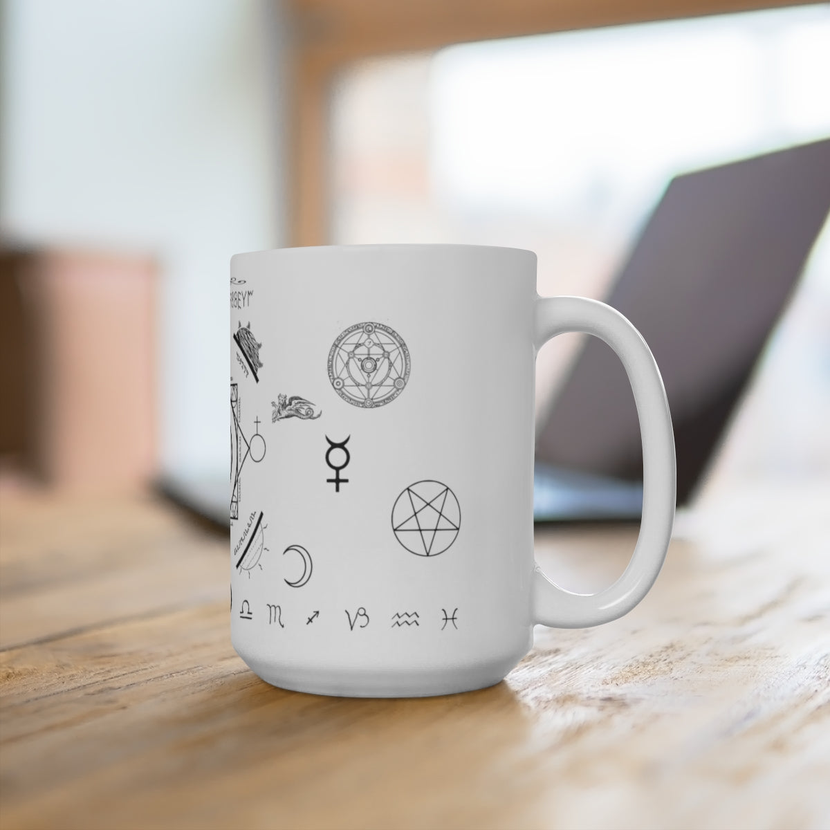 A white mug sitting on a wooden table. On the mug is an assortment of alchemical symbols, including a half moon, a pentacle, the symbol for mercury, zodiac signs along the bottom, etc.