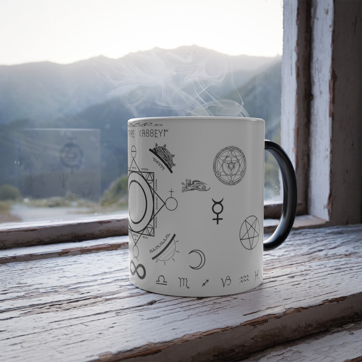 A white mug with a black handle sitting on a windowsill. On the mug is an assortment of alchemical symbols, including a half moon, a pentacle, the symbol for mercury, zodiac signs along the bottom, etc.