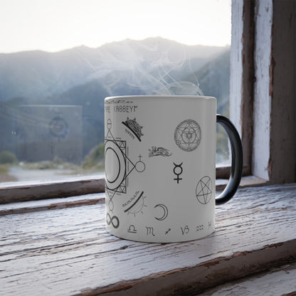 A white mug with a black handle sitting on a windowsill. On the mug is an assortment of alchemical symbols, including a half moon, a pentacle, the symbol for mercury, zodiac signs along the bottom, etc.