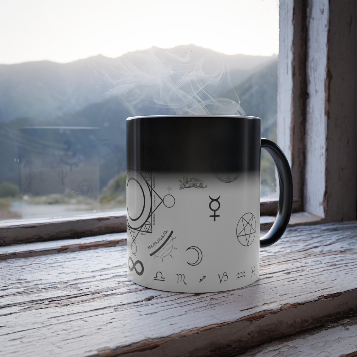 A white mug with a black handle sitting on a windowsill. Part of the mug has changed from white to black as response to the heat of the drink in it. On the mug is an assortment of alchemical symbols, including a half moon, a pentacle, the symbol for mercury, zodiac signs along the bottom, etc.