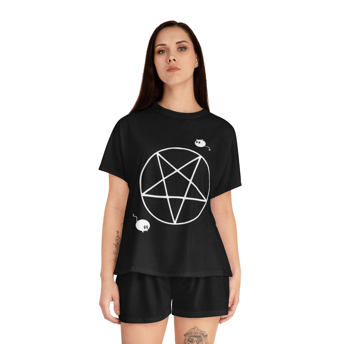 A woman with long black hair and tattoos wearing a black pajama shirt and black pajama shorts. The shirt has an upside down pentacle with a mouse in the bottom left corner and a chibi cat demon in the upper right corner.